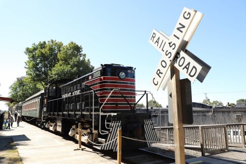 A vintage train at a station next to a railroad crossing sign, surrounded by trees and a wooden platform.