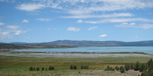 A panoramic view of a serene lake surrounded by green fields and distant mountains under a blue sky with scattered clouds.