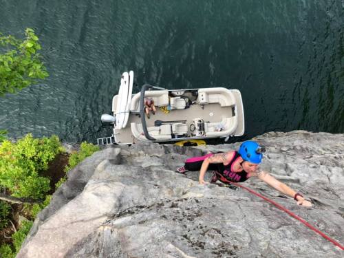 A climber ascends a rocky cliff above a boat on calm water, surrounded by greenery.