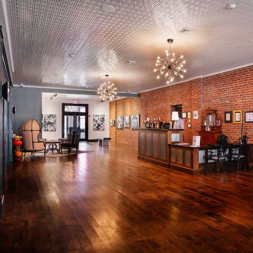 A spacious interior with wooden floors, brick walls, and elegant lighting, featuring seating and art displays.