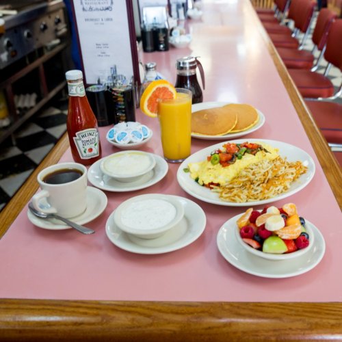A diner table with coffee, orange juice, pancakes, an omelet, and fruit salad on a pink countertop.
