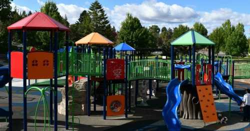 Colorful playground with slides and climbing structures, surrounded by trees and a blue sky with clouds.