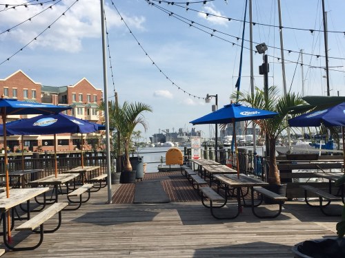 Outdoor dining area with wooden tables, blue umbrellas, and a view of the water and boats in the background.