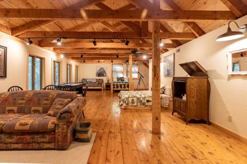 A spacious, rustic living area with wooden beams, a couch, and a bed, featuring large windows and warm lighting.