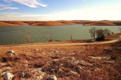 A serene landscape featuring a calm lake surrounded by rolling hills and sparse trees under a clear sky.