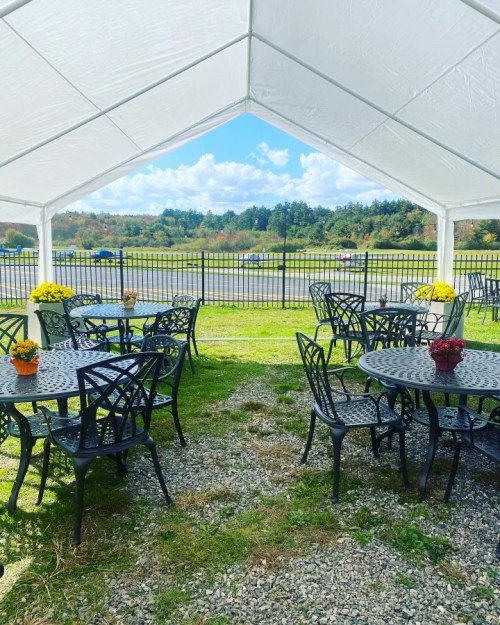 A tented outdoor seating area with black tables and chairs, surrounded by greenery and a view of distant hills.