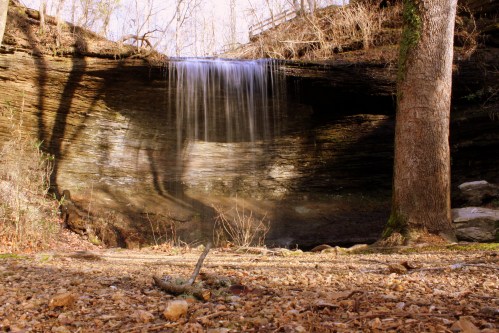 A serene waterfall cascades over rocky cliffs, surrounded by trees and a sandy ground in a tranquil natural setting.