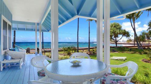 A serene beachfront view from a porch with a round table and chairs, surrounded by palm trees and blue skies.