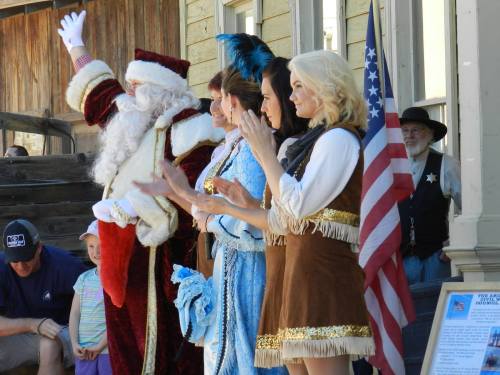 A festive scene with Santa and three women in costumes, clapping and waving, with an American flag in the background.