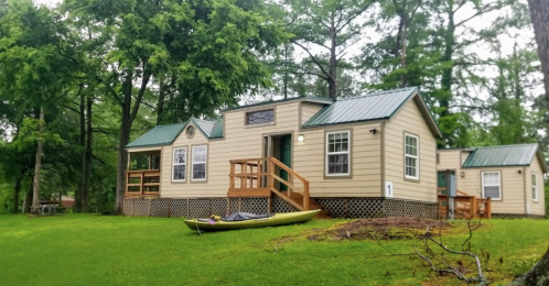 A cozy cabin with a green kayak in front, surrounded by trees and grass on a rainy day.