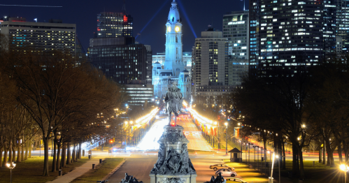 Night view of a city skyline with a clock tower, illuminated streets, and a statue in the foreground.