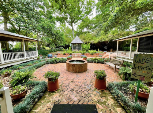 A serene garden with brick pathways, benches, and potted plants, featuring a central fountain and shaded areas.
