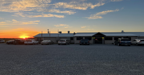 A building at sunset, with a gravel parking lot filled with cars and a colorful sky in the background.