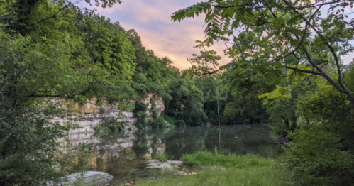 A serene landscape featuring a calm lake surrounded by lush greenery and rocky cliffs under a colorful sky.