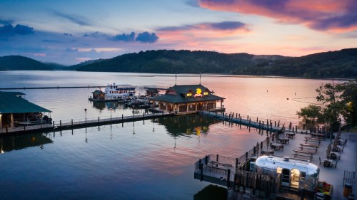 A scenic view of a lakeside restaurant at sunset, with boats docked and calm waters reflecting the colorful sky.