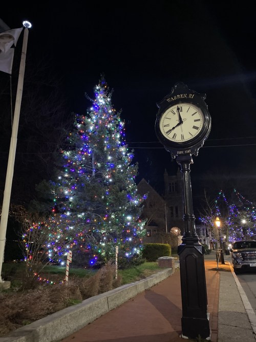 A decorated Christmas tree and a vintage clock in a festive, illuminated town square at night.