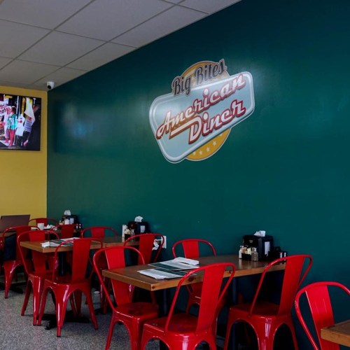 Interior of a diner with red chairs and tables, featuring a green wall and a sign that reads "Big Bites American Diner."