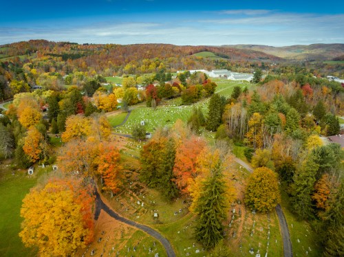 Aerial view of a cemetery surrounded by vibrant autumn trees and rolling hills under a clear blue sky.