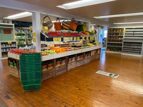 A bright grocery store interior featuring fresh produce, jars, and baskets on display with wooden flooring.