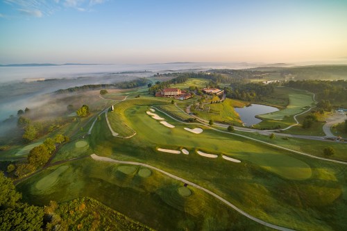 Aerial view of a golf course at sunrise, featuring lush greens, a pond, and misty hills in the background.