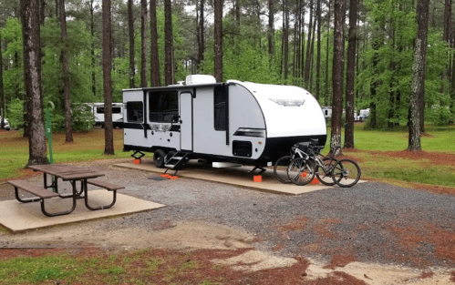 A white travel trailer parked at a campsite surrounded by tall trees, with a picnic table and two bicycles nearby.