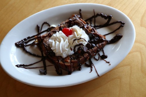A slice of chocolate dessert topped with whipped cream, chocolate chips, and a cherry, served on a white plate.