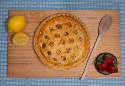 A golden pie with star-shaped cutouts, surrounded by lemons and strawberries on a wooden board.