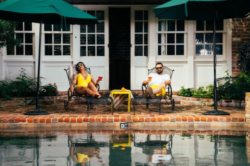 A man and woman relax by a pool, sipping drinks under green umbrellas, with a charming house in the background.