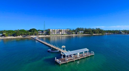 Aerial view of a serene waterfront with a long pier, clear blue water, and a building in the background under a bright sky.