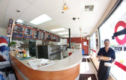 A smiling man holds a pizza in a bright, casual restaurant with a menu board and counter in the background.