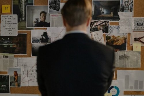 A person in a suit stands in front of a bulletin board filled with photos, notes, and newspaper clippings.