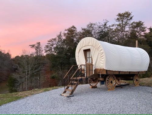 A covered wagon on a gravel area, surrounded by trees, with a colorful sunset sky in the background.