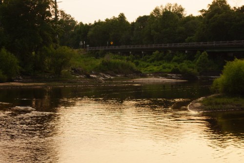 A serene river scene at sunset, with lush greenery and a bridge in the background reflecting on the water's surface.