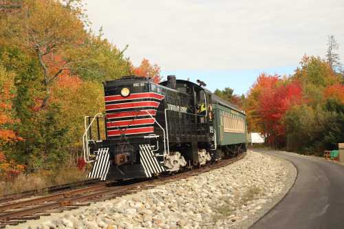 A vintage train with a black and red locomotive and green passenger car travels along a scenic, autumn-colored landscape.