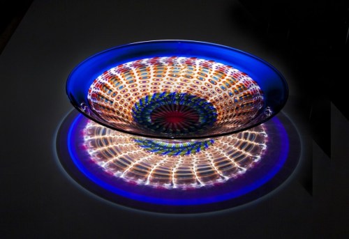 A colorful glass bowl with intricate patterns, casting vibrant reflections on a surface below.