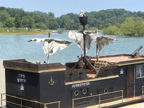 A pirate-themed boat named "The Booty" with tattered sails, docked by a serene lake surrounded by greenery.
