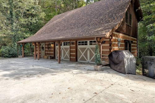 A rustic wooden garage with a sloped roof, surrounded by trees, featuring two garage doors and a concrete driveway.