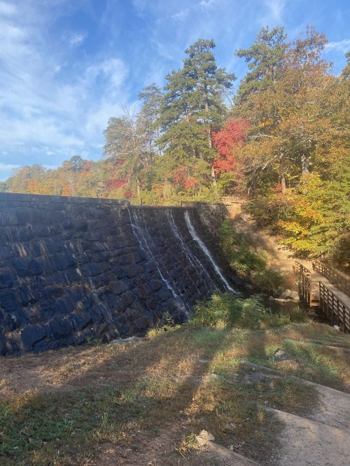 A stone dam with water cascading down, surrounded by trees displaying autumn colors under a clear blue sky.