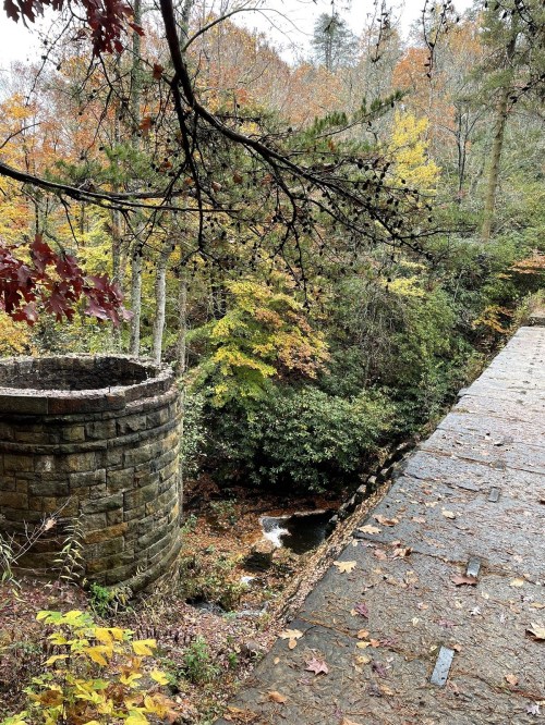 A stone structure beside a winding stream, surrounded by vibrant autumn foliage in a wooded area.