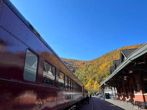 A vintage train alongside a station, with colorful autumn foliage on the hills under a clear blue sky.