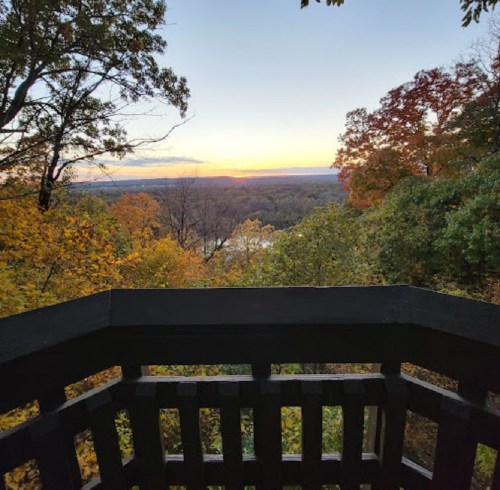View from a wooden balcony overlooking a river and colorful autumn trees at sunset.