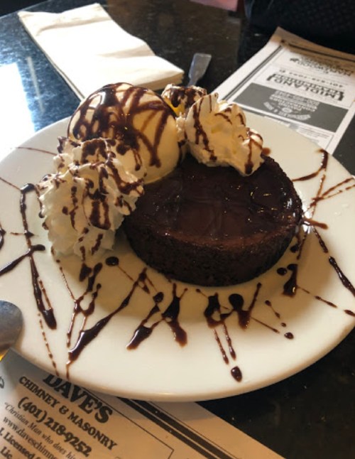 A rich chocolate dessert topped with ice cream and whipped cream, drizzled with chocolate sauce on a white plate.