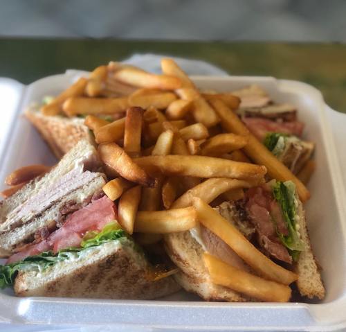 A sandwich with layers of turkey, ham, and lettuce, topped with French fries in a takeout container.