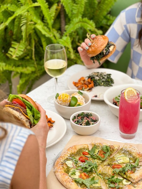 A table with a variety of dishes, including burgers, salads, and drinks, surrounded by greenery.
