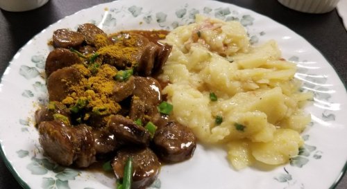 A plate featuring sliced meat in a brown sauce with spices, alongside a serving of creamy mashed potatoes.