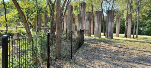 A fenced area with tall, weathered wooden pillars surrounded by trees and grass in a sunny outdoor setting.