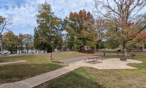 A park scene with trees, a picnic table, and a view of a lake in the background under a partly cloudy sky.