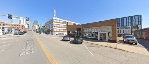 Street view of a city block featuring the RAYGUN store, with cars parked and buildings in the background under a clear sky.
