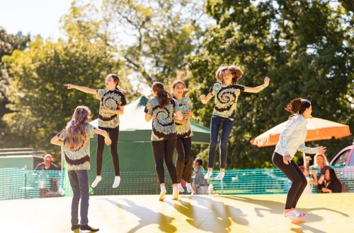 A group of girls in tie-dye shirts joyfully jumping on a large inflatable surface in a sunny park.