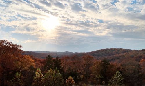 A scenic view of rolling hills covered in autumn foliage under a partly cloudy sky with the sun shining.